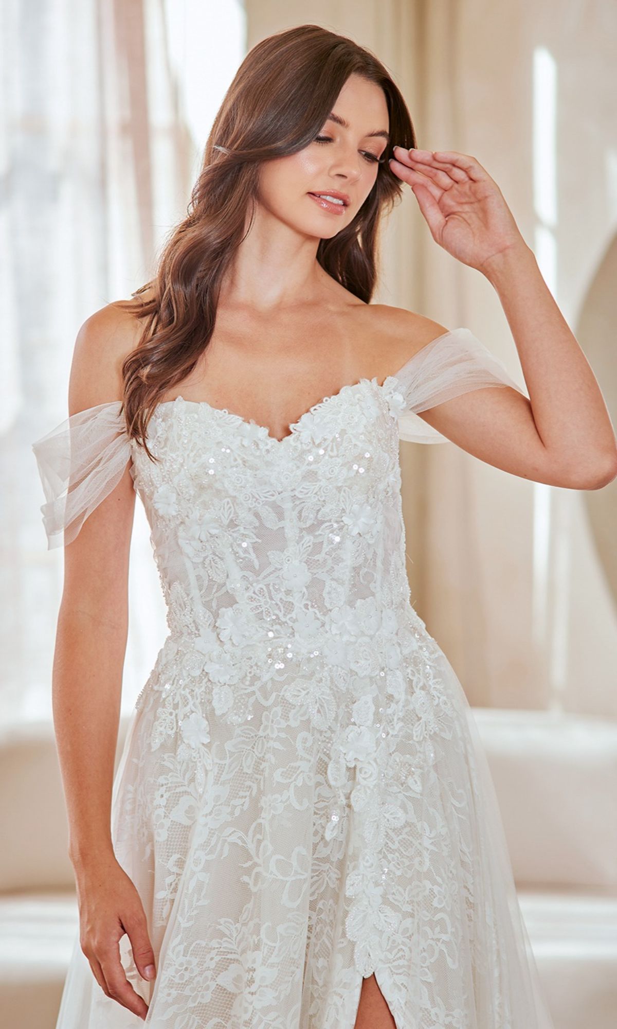 A woman stands indoors looking down, wearing the Amelia Couture 2139 long wedding dress—an off-the-shoulder lace gown with floral appliqué—and raises one hand near her face.